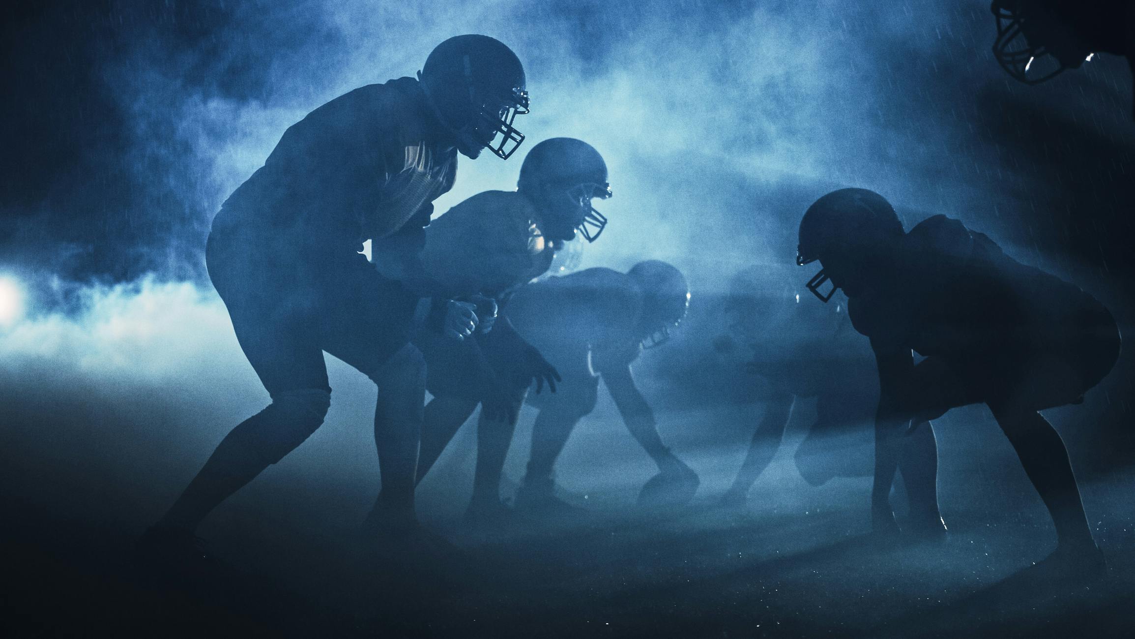 American Football Field Two Teams Compete: Players Pass, Run, Attack to Score Touchdown Points. Rainy Night with Athletes Fight for the Ball in Dramatic Smoke.