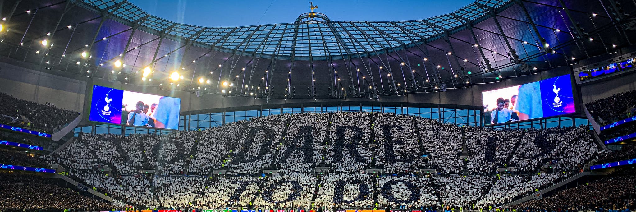 Jalkapallostadion täynnä yleisöä, joka muodostaa koreografian pitämällä ylhäällä valkoisia ja tummia papereita. Katsomossa erottuu suuri teksti, ja kentän yläpuolella näkyy jättiscreenit sekä kirkkaat valot stadionin katossa