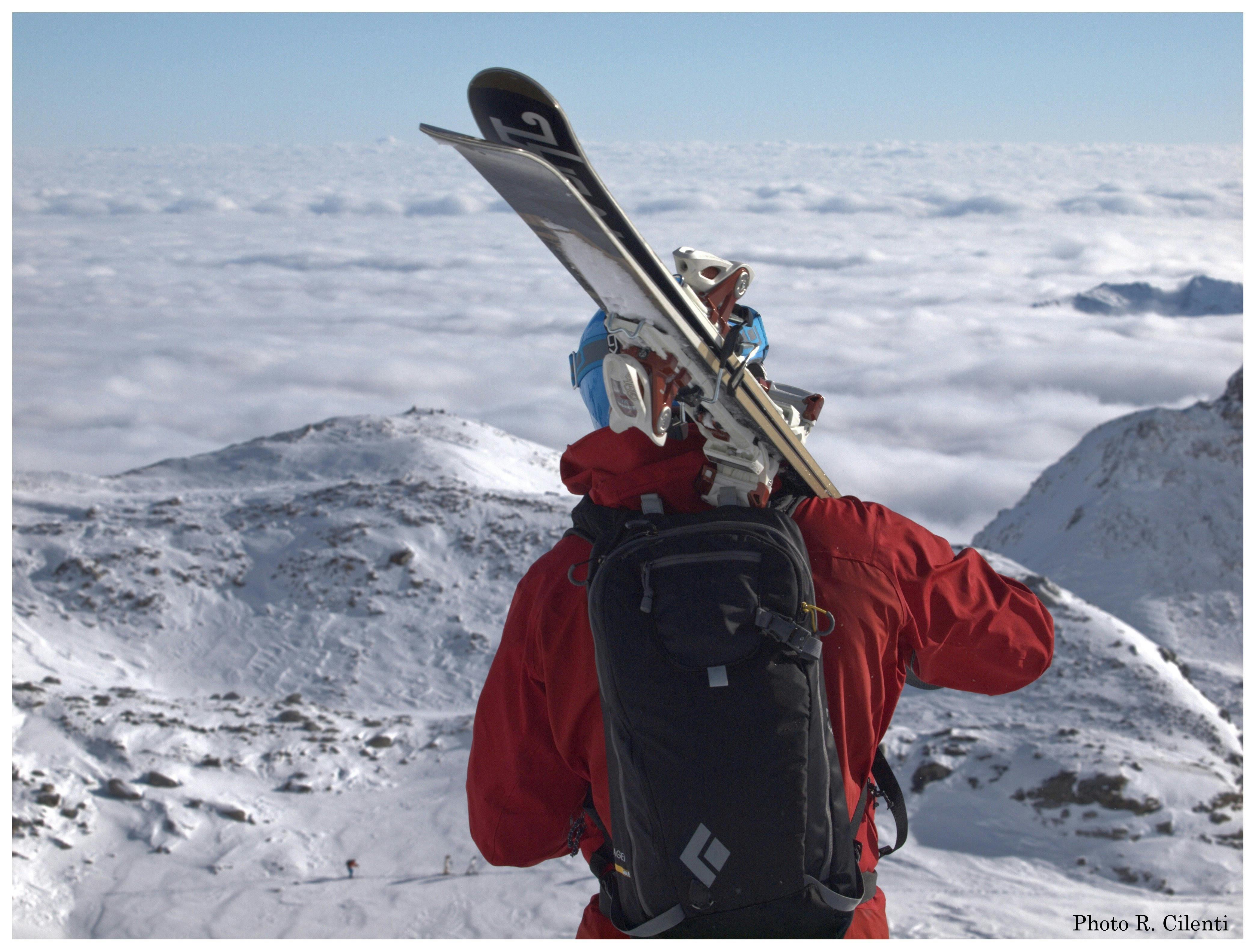 Laskettelijasta lähikuva, jossa hän on selkä kameraan päin. Olalla laskettelusukset ja taustalla pilvinen vuoristomaisema.
