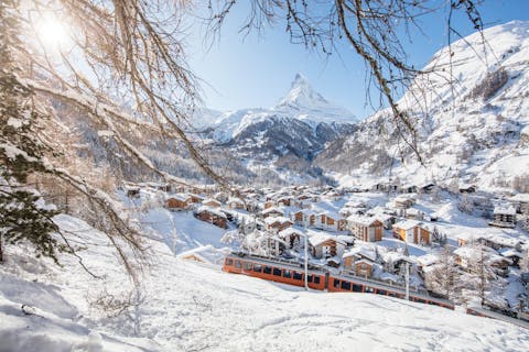 Zermattin kylää kuvattu ylhäältä päin. Kylän edustalla kulkee juna ja taustalla luminen vuoristo.