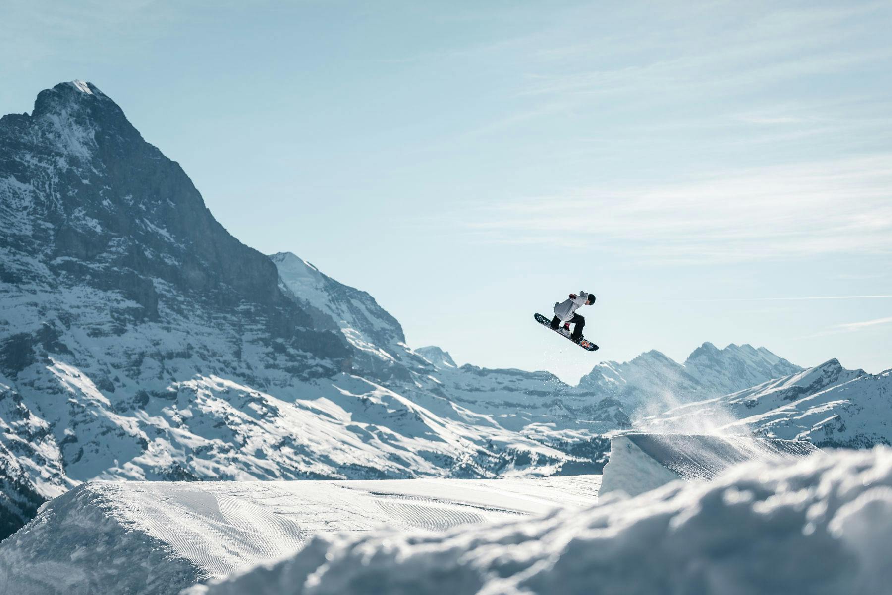 Lumilautailija hyppäämässä hyppyristä. Taustalla vuoristo.