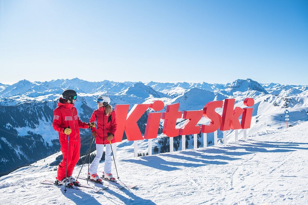 Pariskunta KitzSki logon edessä fiilistelemässä vuoren huipulla lasketteluvälineet alla.