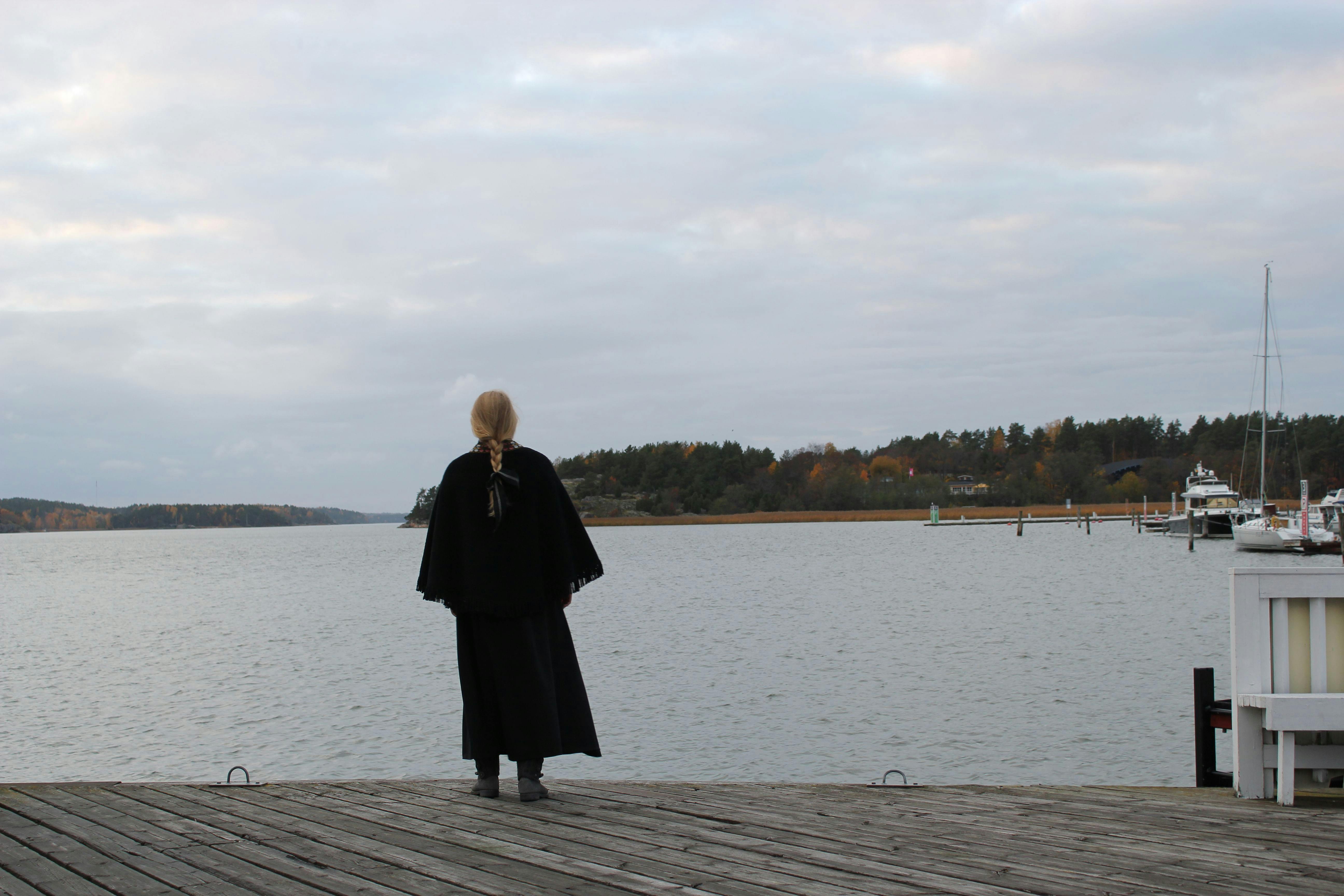 Nainen laiturilla pukeutuneena mustaan kaapuun, samalla katsoen merimaisemaa.