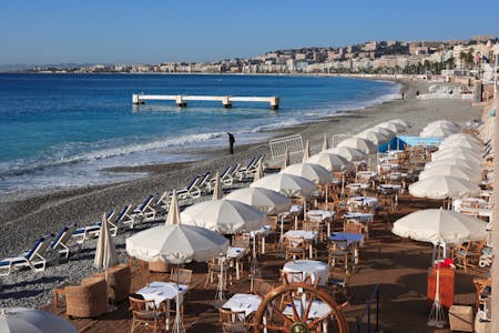 Rantanäkymä Nizzan rannasta, jossa on terassi täynnä valkoisia aurinkovarjoja sekä ruokaryhmiä.